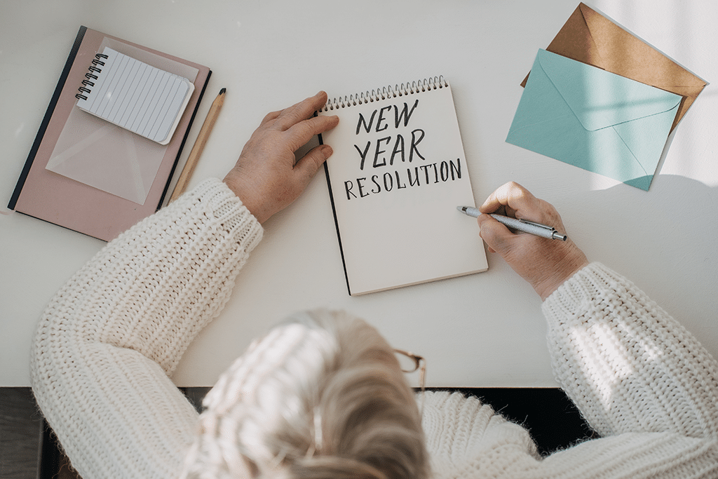 woman in white sweater writing handwritten text New Years resolutions in open notepad on the table